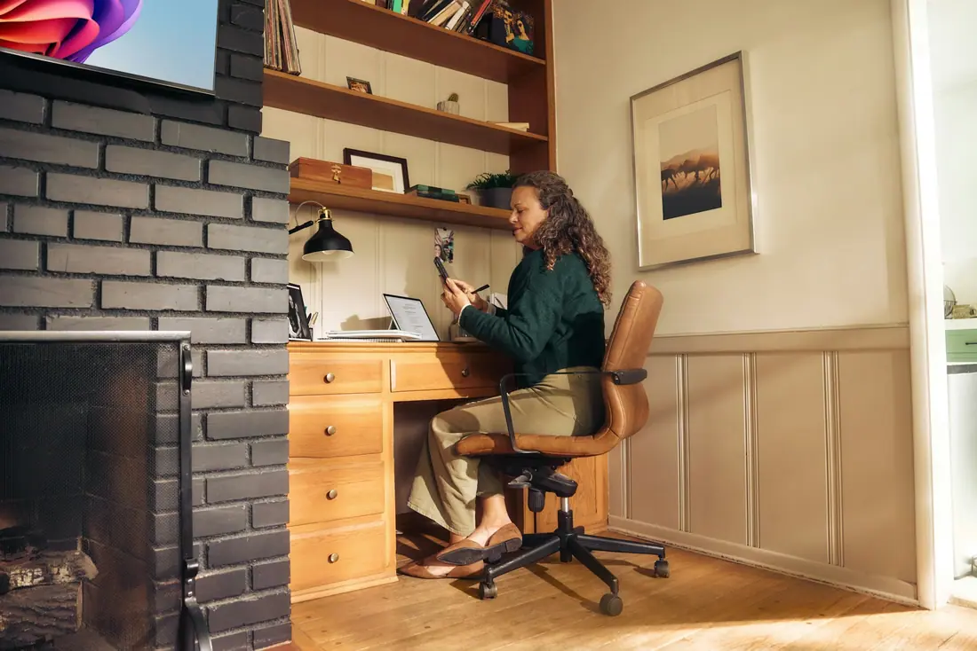 Person at a wooden desk in a cozy home office with a laptop and phone