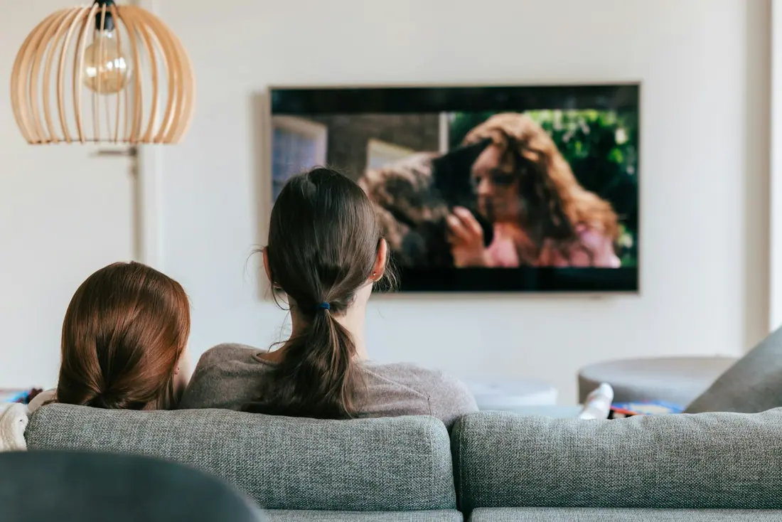 Two people watching a show on a wall-mounted television from a grey sofa